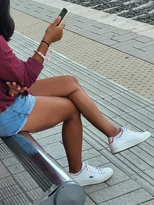 Candid Girl in Denim Shorts And White Sneakers Sitting With Crossed Legs on the Bus Stop Street Creepshot