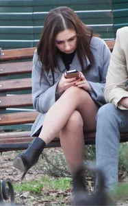 Nice candid girl with crossed legs in pantyhose sitting on a park bench reading something on her phone