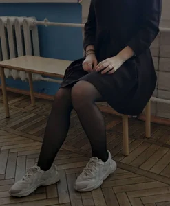College teen wearing a black dress, pantyhose and sneakers gently touching her thigh while sitting on a bench in a hallway